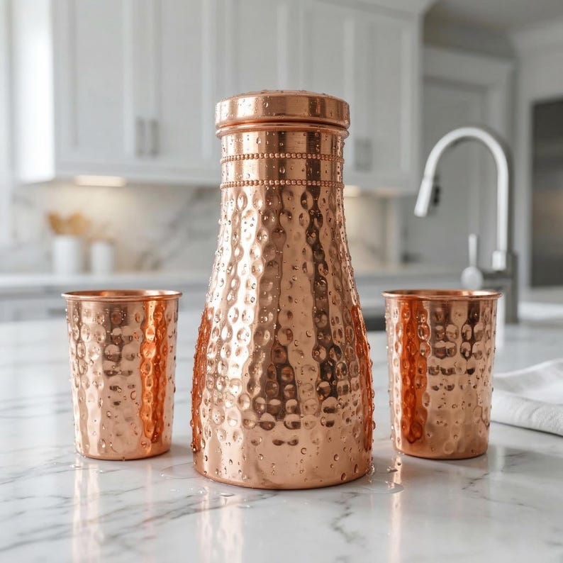 May include: A copper water pitcher with a hammered texture, a matching lid, and two copper cups. The pitcher and cups are displayed on a marble countertop. The kitchen background includes white cabinets and a stainless steel faucet.