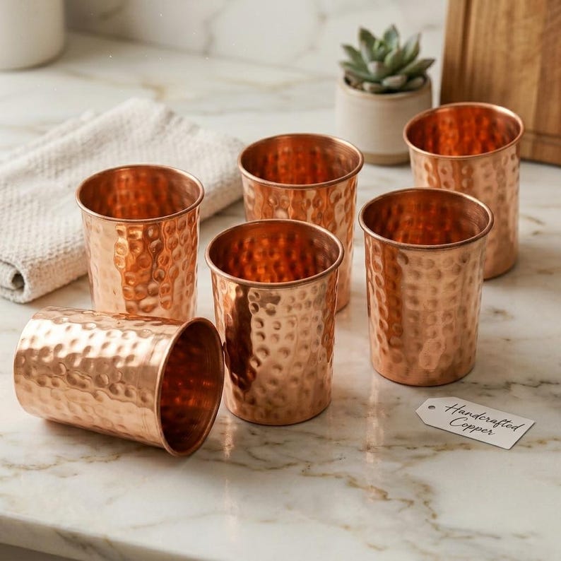 Puede incluir: Seis vasos de color cobre con textura martillada. Un vaso est&aacute; tumbado de lado. Una peque&ntilde;a etiqueta dice "Handcrafted Copper". Los vasos est&aacute;n colocados sobre una superficie de m&aacute;rmol.