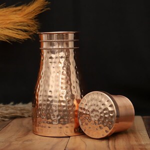 May include: A copper-colored water pitcher with a hammered texture and a matching lid. The pitcher has a wide base that tapers to a narrower neck, with decorative bands near the top. The lid is round and sits beside the pitcher.