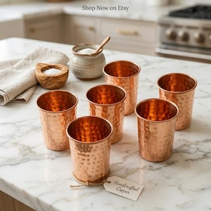 Puede incluir: Seis vasos de cobre con textura martillada est&aacute;n dispuestos sobre una superficie de m&aacute;rmol. Los vasos son de color cobre c&aacute;lido y tienen forma cil&iacute;ndrica. Una etiqueta que dice "Handcrafted Copper" est&aacute; adherida a uno de los vasos. El texto "Shop Now on Etsy" es visible.