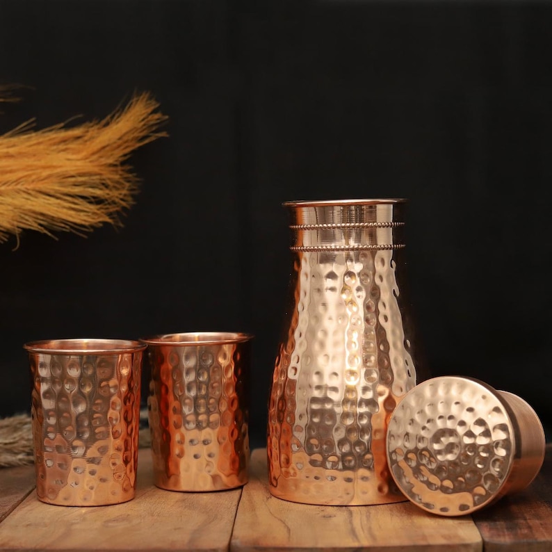 May include: A copper pitcher, two drinking glasses, and a lid with a hammered texture. The copper items are displayed on a wooden surface against a dark background. The pitcher has a narrow neck and a wide base.