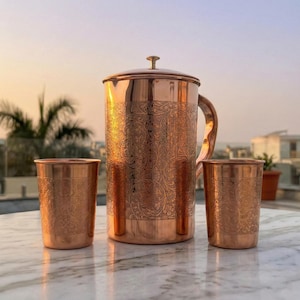 Puede incluir: Una jarra de cobre con tapa y asa, acompa&ntilde;ada de dos vasos de cobre a juego. Cada pieza presenta un intrincado patr&oacute;n floral en relieve que cubre la superficie exterior, reflejando un tono marr&oacute;n rojizo met&aacute;lico y c&aacute;lido.