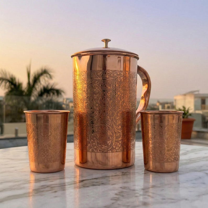 Puede incluir: Una jarra de cobre con tapa y asa, acompa&ntilde;ada de dos vasos de cobre a juego. Cada pieza presenta un intrincado patr&oacute;n floral en relieve que cubre la superficie exterior, reflejando un tono marr&oacute;n rojizo met&aacute;lico y c&aacute;lido.