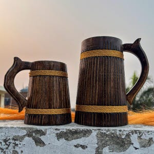 May include: Two wooden beer tankards with carved handles and vertical grooves. The dark brown tankards are wrapped with a gold-coloured rope around the middle. The tankards are of different sizes, with the larger one on the right.