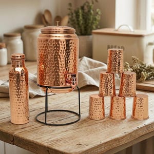 Puede incluir: Dispensador de agua de cobre con textura martillada, una botella a juego y un juego de tazas de cobre. El dispensador se encuentra sobre un soporte de metal negro. Los art&iacute;culos de cobre tienen un tono c&aacute;lido marr&oacute;n rojizo.