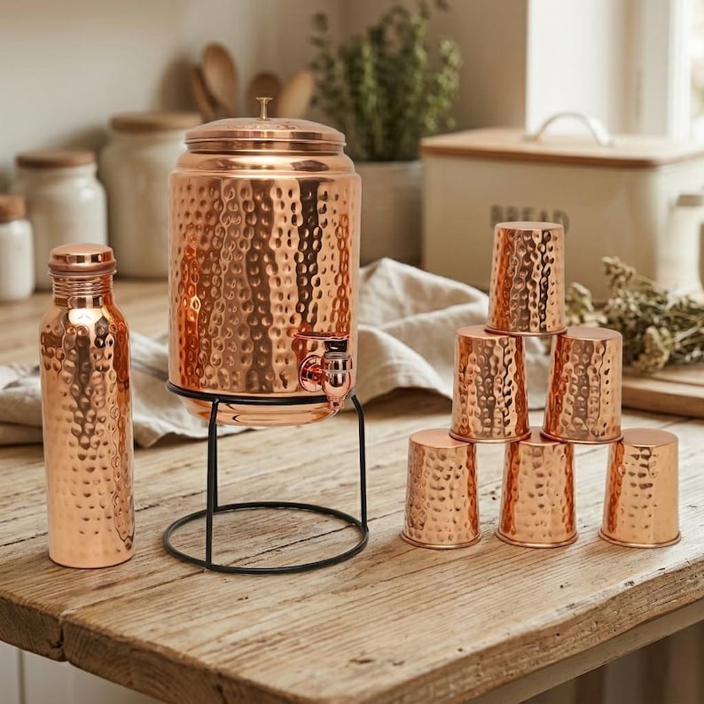 Puede incluir: Dispensador de agua de cobre con textura martillada, una botella a juego y un juego de tazas de cobre. El dispensador se encuentra sobre un soporte de metal negro. Los art&iacute;culos de cobre tienen un tono c&aacute;lido marr&oacute;n rojizo.