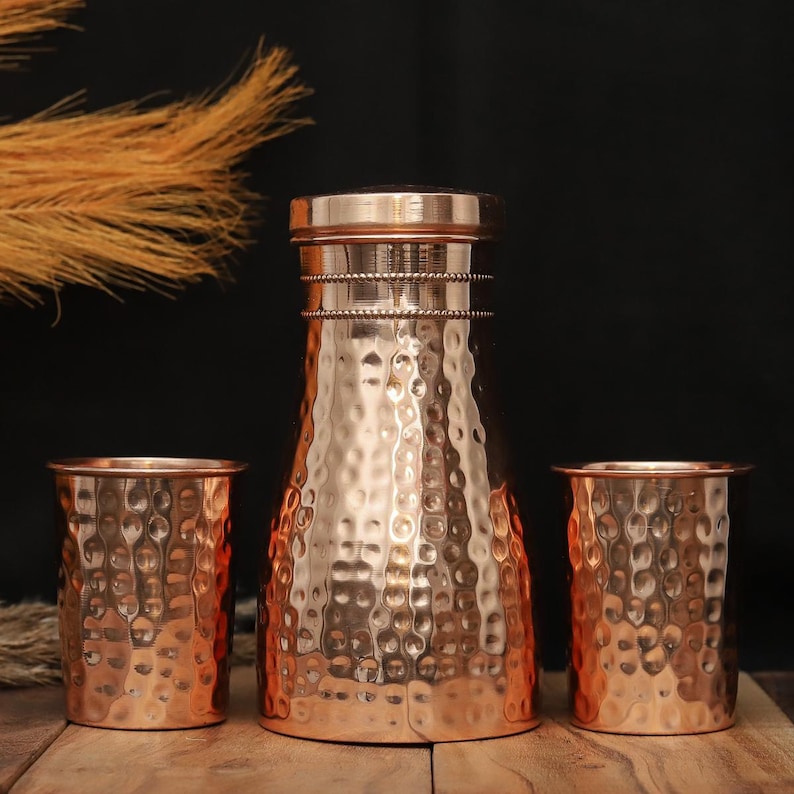 May include: A copper water pitcher and two matching tumblers with a hammered texture. The pitcher has a rounded body and a lid. The tumblers are cylindrical. The set is displayed on a wooden surface against a dark background.