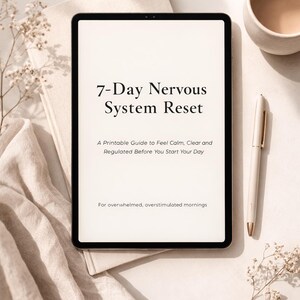 May include: A digital tablet displays the text "7-Day Nervous System Reset" with additional text below. A pen, a cup of coffee, and a book are also on the white surface. The image has a neutral, calming aesthetic.