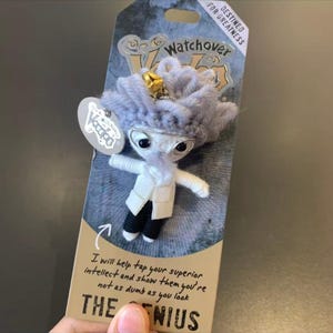 May include: A Watchover Voodoo doll, The Genius, with a gray yarn hair, glasses, and a white coat. The doll is attached to a card with the text "DESTINED FOR GREATNESS" and "I will help tap your superior intellect and show them you're not as dumb as you look."