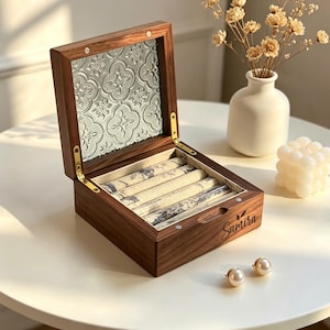 May include: A wooden jewelry box with a hinged lid, featuring a decorative glass panel and a patterned interior. The box is open, revealing several cushioned compartments. Two pearl earrings are placed in front of the box.