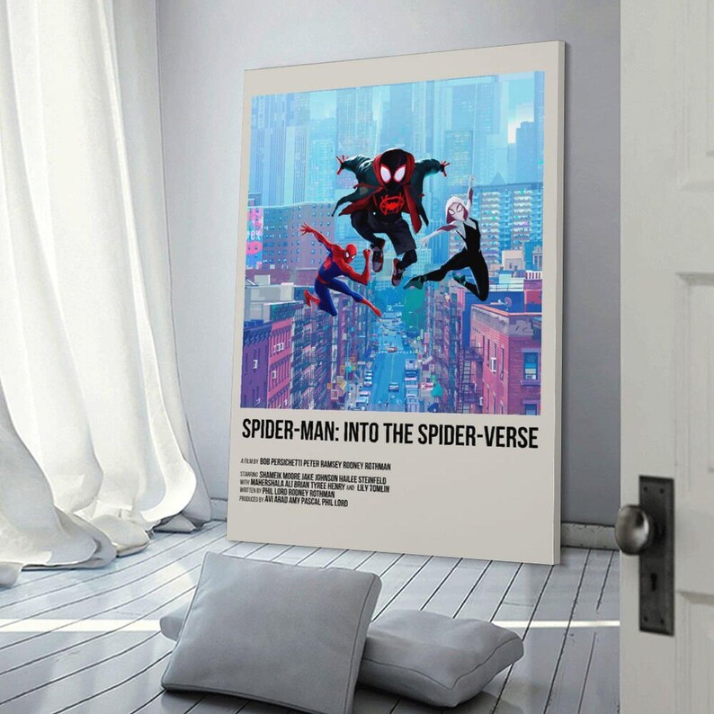 May include: A framed poster featuring the title "Spider-Man: Into the Spider-Verse." The artwork depicts three Spider-Man characters mid-air above a cityscape. The poster is set against a white wall with a window and pillows in the foreground.