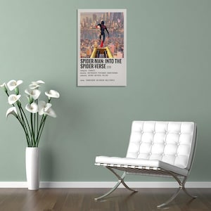 May include: A framed movie poster for "Spider-Man: Into the Spider-Verse" (2018) featuring a cityscape and a Spider-Man figure. The poster is displayed on a light green wall, next to a white vase with white flowers and a white chair.