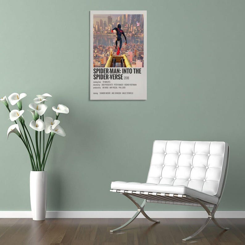 May include: A framed movie poster for "Spider-Man: Into the Spider-Verse" (2018) featuring a cityscape and a Spider-Man figure. The poster is displayed on a light green wall, next to a white vase with white flowers and a white chair.