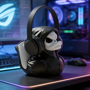 Pode incluir: Um pato de borracha preto usando uma capa preta com capuz e fones de ouvido. O pato tem um rosto branco com um design de caveira. O fundo inclui um monitor de computador e componentes, sugerindo um tema de jogos.