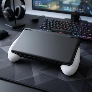 Pode incluir: Consola de jogos portátil preta com punhos brancos, pousada num tapete de rato geométrico. Um monitor de computador, um teclado e auscultadores estão no fundo, sugerindo uma configuração de jogos.