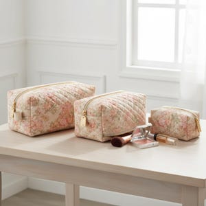 May include: Three quilted cosmetic bags in different sizes, patterned with pink and cream florals. Each bag has a gold zip and a matching gold zip pull. Makeup brushes and compacts are also on the table.