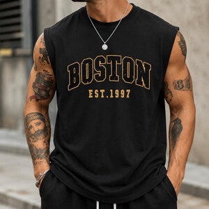 May include: A black sleeveless shirt featuring the word BOSTON in a large, arched, distressed gold font with a black outline, positioned above the text EST. 1997 in a smaller, matching gold font. The shirt has a crew neckline and wide armholes.