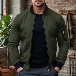 May include: An olive green bomber jacket with a black collar and cuffs. The jacket has a zipper closure and a zippered pocket on the left sleeve. The person is wearing a black shirt and gray pants.