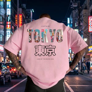 May include: A light pink t-shirt with the word "TOKYO" in a colorful, cityscape design. Below the word "TOKYO" is Japanese text and the phrase "LAND OF THE RISING SUN". The word "JAPAN" is above "TOKYO".