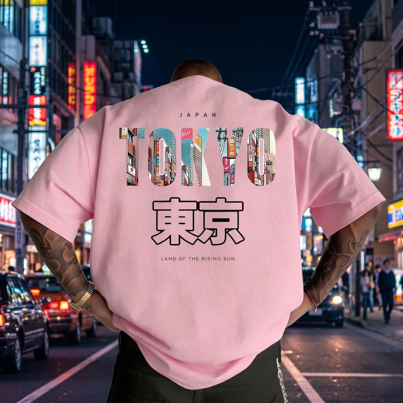May include: A light pink t-shirt with the word "TOKYO" in a colorful, cityscape design. Below the word "TOKYO" is Japanese text and the phrase "LAND OF THE RISING SUN". The word "JAPAN" is above "TOKYO".