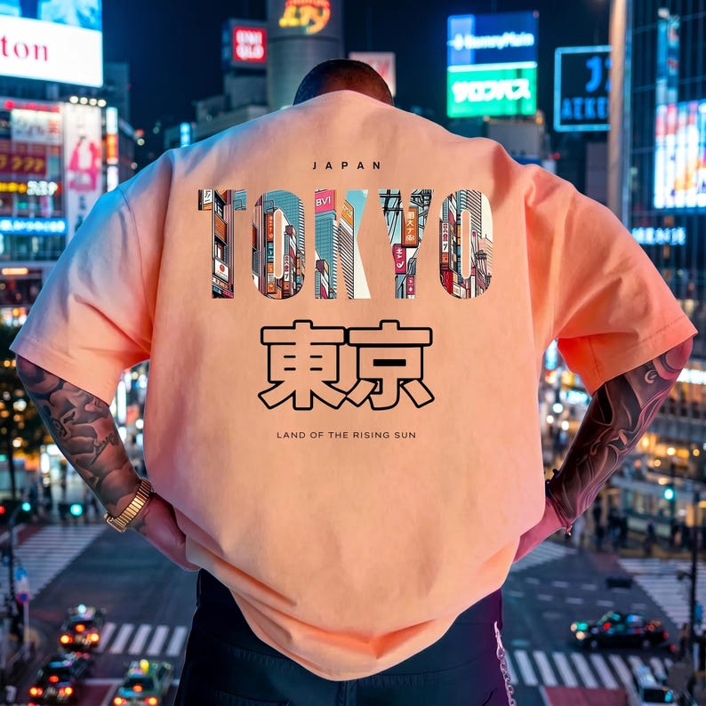 May include: Peach-colored t-shirt with the word "TOKYO" in a cityscape design, "JAPAN" above, and Japanese characters and "LAND OF THE RISING SUN" below. The shirt is worn in front of a city at night.