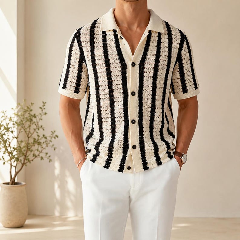 May include: A short-sleeved, button-down shirt with a cream-colored base and vertical black stripes. The shirt has a classic collar and is paired with white pants. The style is casual and suitable for warm weather.