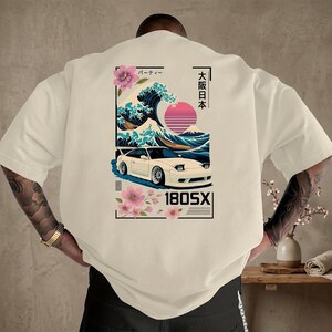 May include: Off-white t-shirt with a graphic of a white sports car set against a Japanese wave and mountain backdrop. Pink flowers and Japanese text surround the car. The text "180SX" is below the car.
