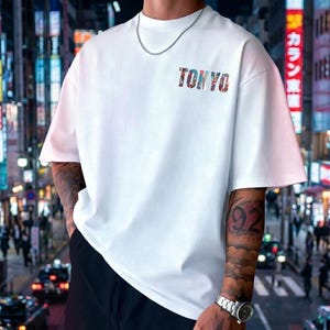 May include: White short-sleeve t-shirt with the word "TOKYO" printed across the chest in a colorful, patterned font. The shirt has a relaxed fit and is worn with a silver chain necklace.