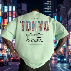 May include: A light green t-shirt with the word "TOKYO" in large, colorful letters, with the Japanese characters for Tokyo below. The shirt also includes the text "JAPAN" and "LAND OF THE RISING SUN". The design features a cityscape.