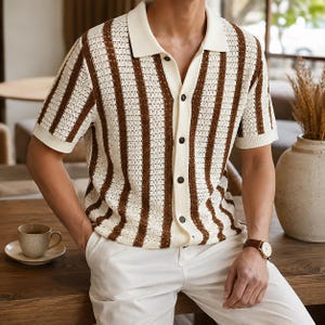 May include: A short-sleeved, button-down shirt with a cream-colored collar and vertical brown stripes. The shirt is paired with white pants. A wristwatch with a brown leather band is visible.