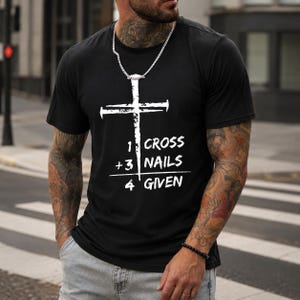 Puede incluir: Una camiseta negra de manga corta con un gráfico blanco de una cruz hecha con un clavo. El texto dice 1 CROSS + 3 NAILS = 4 GIVEN. El diseño está centrado en el pecho. La camiseta tiene cuello redondo y un corte estándar hecho de algodón.