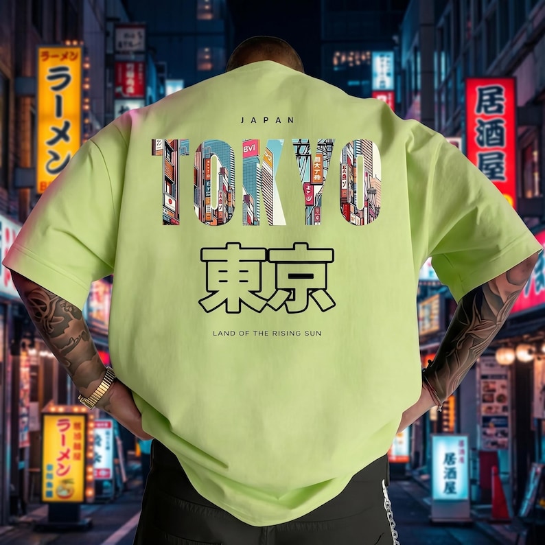 May include: A light green t-shirt with the word "TOKYO" in a colorful, cityscape design. Below, Japanese characters and the text "LAND OF THE RISING SUN" are printed. The word "JAPAN" is above the word "TOKYO".