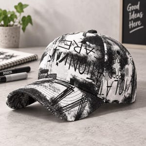 Puede incluir: Una gorra de béisbol con un estampado abstracto en blanco y negro, texturas de tinta desgastadas y texto manuscrito disperso que incluye las palabras "VIOLATION" y "CARE". La gorra tiene una visera curva y una corona estructurada.