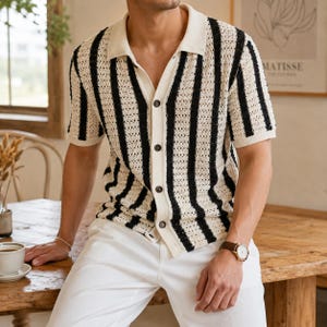 May include: A short-sleeved, button-down shirt with a cream-colored base and vertical black stripes. The shirt has a collared neckline and is paired with white pants. The shirt's design is a knit pattern.