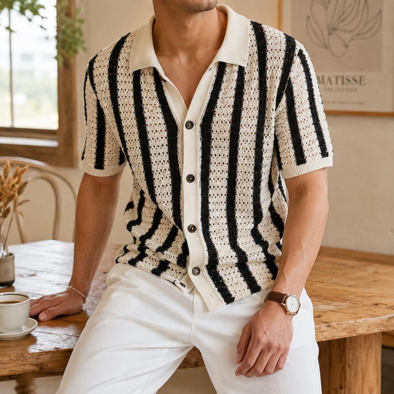 May include: A short-sleeved, button-down shirt with a cream-colored base and vertical black stripes. The shirt has a collared neckline and is paired with white pants. The shirt's design is a knit pattern.