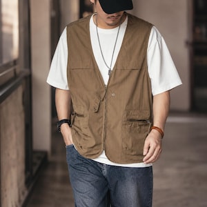 May include: A brown utility vest with multiple pockets and a zip-up front. The vest is worn over a white t-shirt and paired with blue jeans. The vest has a casual, functional design.