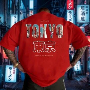 May include: A red t-shirt with the word "TOKYO" in a multi-colored, cityscape-filled font. Below the word "TOKYO" is Japanese text and the phrase "LAND OF THE RISING SUN". The word "JAPAN" is above "TOKYO".