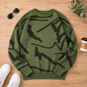 Puede incluir: Un suéter de manga larga y cuello redondo en color verde oliva con un patrón abstracto en verde oscuro. La prenda de punto tiene cuello, puños y dobladillo acanalados. El diseño consiste en líneas oscuras irregulares y curvas.