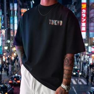 May include: Black t-shirt with the word "TOKYO" in colorful, patterned letters. The shirt has short sleeves and a crew neck. The background shows a city street at night with bright lights.