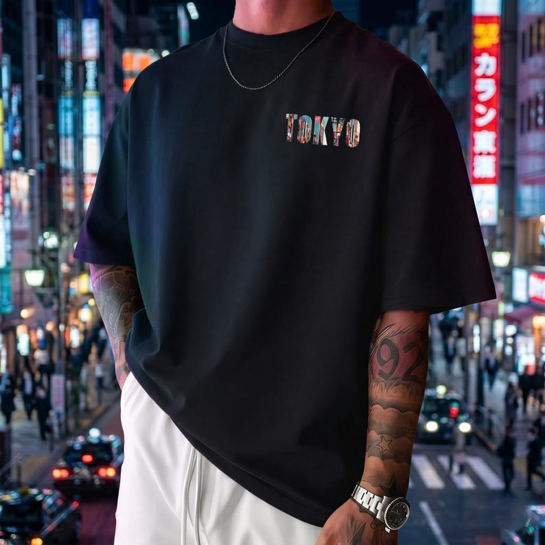 May include: Black t-shirt with the word "TOKYO" in colorful, patterned letters. The shirt has short sleeves and a crew neck. The background shows a city street at night with bright lights.