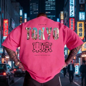 May include: A pink t-shirt with the word "TOKYO" in a cityscape design, and Japanese characters below. The text "JAPAN" is above the word "TOKYO" and "LAND OF THE RISING SUN" is below. The background shows a city street at night.