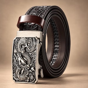 Puede incluir: Un cinturón de cuero marrón oscuro con una hebilla rectangular de metal plateado que presenta un diseño de dragón en relieve. La correa tiene un patrón intrincado y repetitivo grabado a lo largo y una trabilla de cuero liso.