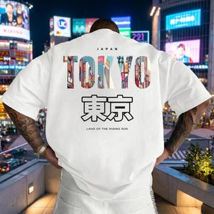 May include: White t-shirt with a graphic design featuring the word "TOKYO" in colorful letters, with Japanese characters below. The text "JAPAN" is above the word "TOKYO" and "LAND OF THE RISING SUN" is below.