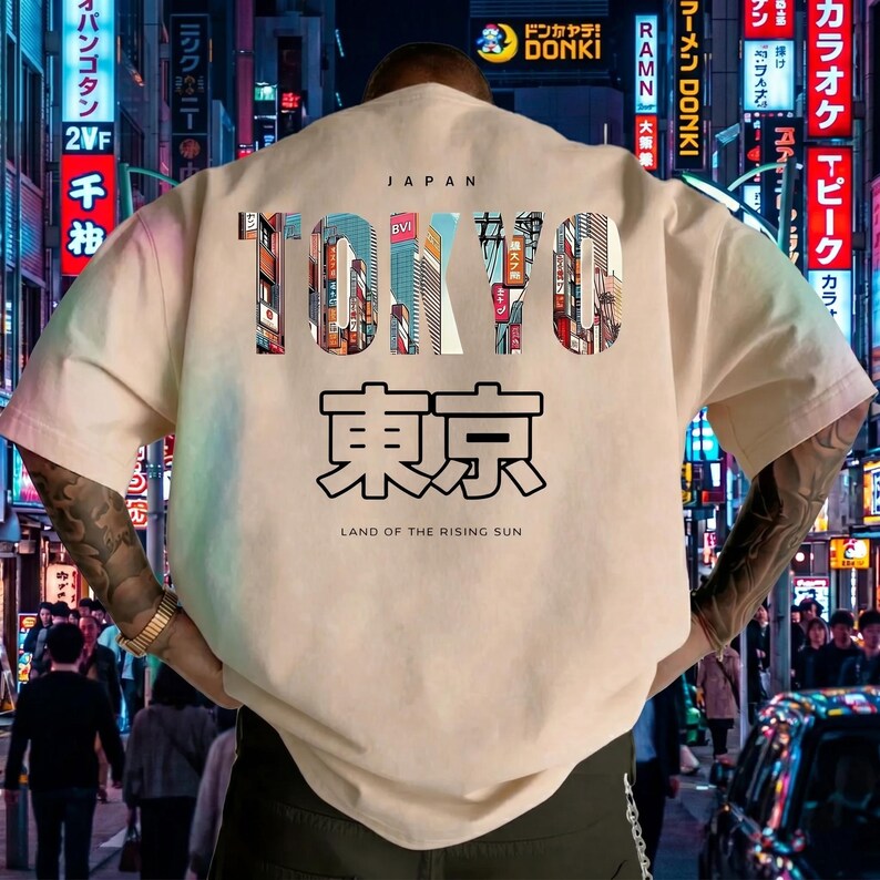 May include: Cream-colored t-shirt with a Tokyo-themed design. The word "TOKYO" is displayed in a colorful, cityscape-filled font, with Japanese characters below. The text "LAND OF THE RISING SUN" is printed at the bottom.