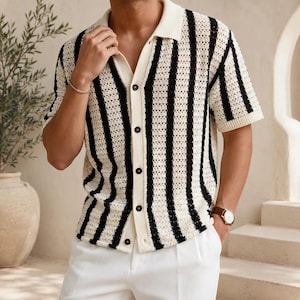 May include: A short-sleeved, button-down shirt with a cream-colored base and bold black vertical stripes. The shirt features a classic collar and is paired with white pants. The knit fabric has a textured appearance, and the shirt has dark buttons.