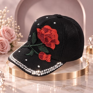May include: A black baseball cap featuring an embroidered red rose with green leaves on the front panel. The cap is decorated with scattered clear rhinestones across the crown and two rows of rhinestones lining the edge of the curved brim.