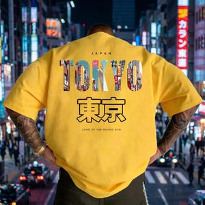 May include: A yellow t-shirt with the word "TOKYO" in a colorful, cityscape-filled font. Below, Japanese characters and the phrase "LAND OF THE RISING SUN" are printed. The shirt is a casual style.