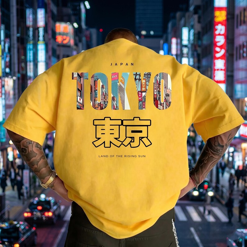 May include: A yellow t-shirt with the word "TOKYO" in a colorful, cityscape-filled font. Below, Japanese characters and the phrase "LAND OF THE RISING SUN" are printed. The shirt is a casual style.