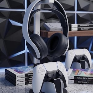 May include: A white and black gaming headset and controller stand. The stand holds a black and white headset and a white controller. The base is black. The image also shows a controller, game cases, and a console.
