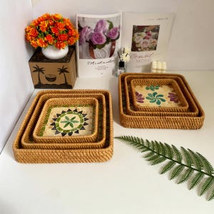 Puede incluir: Juego de tres bandejas cuadradas de ratán tejido. Las bandejas están anidadas, con un diseño de mosaico en el centro de la bandeja más pequeña. El mosaico presenta colores verdes, marrones y beige. Las bandejas exteriores son de color marrón natural.